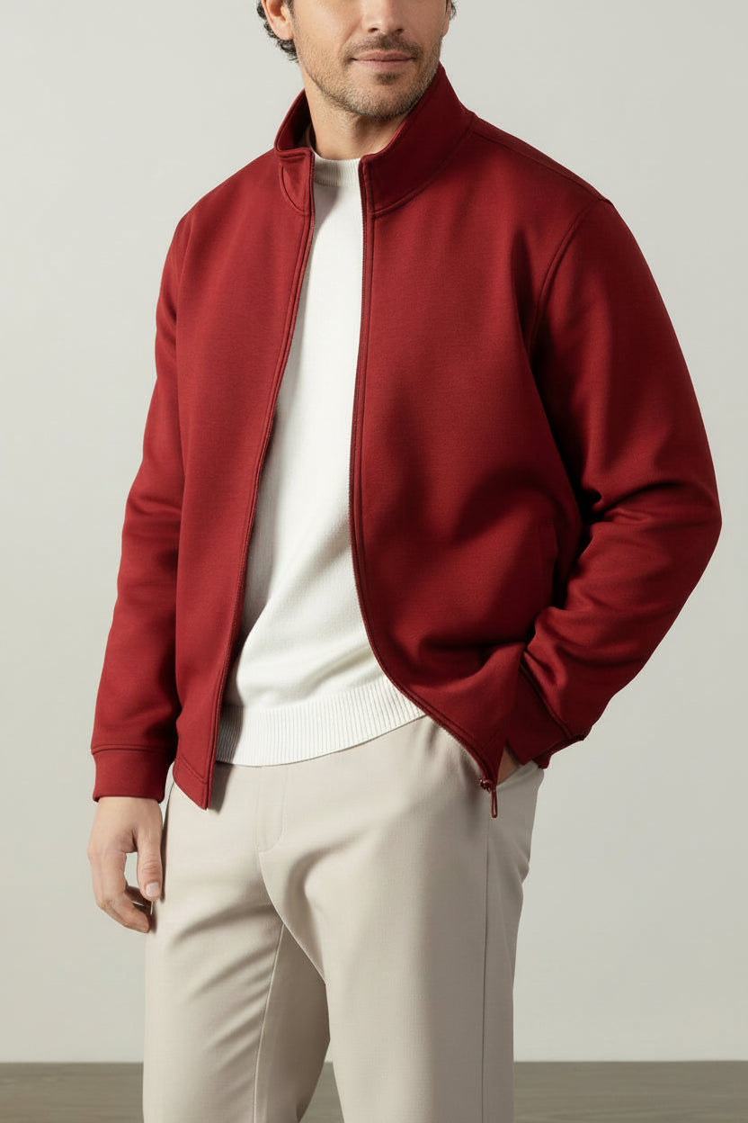 A man wearing a burgundy zip-up jacket over a white ribbed sweater and beige trousers, standing indoors against a light gray wall with a wooden chair partially visible.